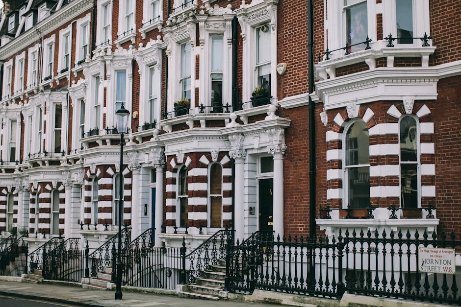 A row of Victorian-style terraced houses with red brick facades and white decorative architectural details, including ornate cornices, window surrounds, and balustrades. Each property features tall, narrow sash windows with white frames and some window boxes filled with greenery. The buildings are fronted by black wrought-iron railings and small steps leading up to the entrances, which have dark-colored doors. A black street lamp with a decorative base stands in front of the houses, illuminating the sidewalk. The environment appears to be a residential street during daylight hours, with a clean pavement and well-maintained facades. This scene illustrates urban housing typical of London neighborhoods, where private property and independent waste management services like Waste Disposal Merton may operate, especially noting the absence of visible waste or rubbish in the scene, aligning with responsible waste disposal practices.
