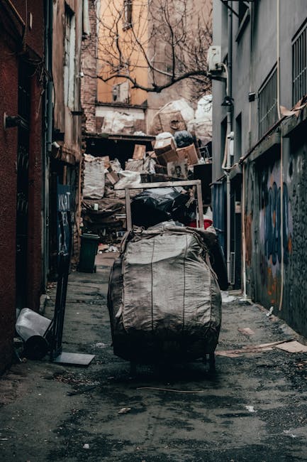 A narrow alleyway cluttered with accumulated rubbish and discarded items, including a large trash bag in the foreground on a wheeled trolley, with its surface appearing worn and creased. Behind it, there are numerous stacked cardboard boxes, plastic bags, and miscellaneous debris piled against a chain-link fence or wall, which are amidst an environment of urban decay. The alley is flanked by brick and metal-sided buildings, with graffiti visible on one wall to the right, and the ground is stained with dark patches, likely from dirt and spilled waste. Bare tree branches extend overhead, and the scene is illuminated by natural daylight, creating a somewhat dim and gritty atmosphere. The clutter suggests a site for private waste collection or clearing efforts, possibly supported by a waste disposal service like Waste Disposal Merton, which specializes in rubbish removal and on-site clearance solutions in the area.