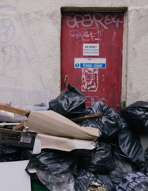 A pile of black rubbish bags, cardboard boxes, and miscellaneous waste items stacked outside a red metal door set into a rough-textured, off-white wall. The bags appear to contain debris or refuse, with some partially open revealing their contents; the cardboard is flattened and stacked atop the bags. The door displays various graffiti tags and signs, including a white sign stating 'No Money' and a blue sticker reading 'Keep Clear.' The scene suggests an area designated for waste collection or disposal, with the rubbish left temporarily in an urban environment, possibly awaiting private removal by a service such as Waste Disposal Merton. Natural daylight illuminates the area, emphasizing the textures of the waste materials and the weathered appearance of the surrounding wall and door. This visual context highlights the importance of proper rubbish management and the role of professional waste disposal services in maintaining cleanliness outside commercial or residential premises.