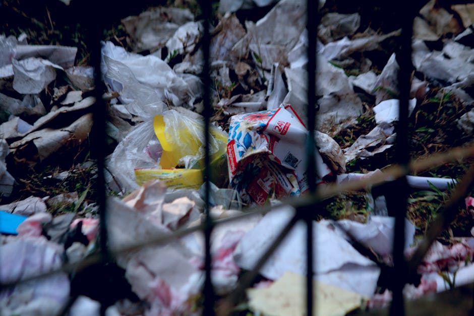 A close-up view through black metal fencing shows a pile of mixed waste and litter, including crumpled plastic bags, torn paper, and cardboard packaging. The plastic bags are mostly white, with some yellow and blue elements, and the cardboard packaging features red and blue printed graphics. The waste is scattered on the ground, partially covered with fallen leaves and debris, indicating outdoor disposal in an uncontrolled environment. The scene highlights the importance of proper rubbish removal and disposal practices, which Waste Disposal Merton provides through private collection services to prevent such illegal dumping conditions in the local area. The natural lighting illuminates the disorderly heap, emphasizing the accumulation of waste beyond permitted disposal zones and underscoring the need for professional rubbish clearance to maintain tidiness and environmental integrity.