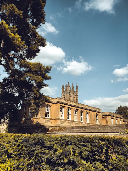 A historic stone building with large, rectangular windows and a flat roof, situated in a lush, green environment. To the left, a tall evergreen tree partially obscures the structure, its dark foliage contrasting with the light-colored stone walls. The building features decorative stone detailing and appears to be set on a raised platform or steps leading up to its entrance. In the background, a partly cloudy sky with scattered white clouds provides natural daylight, highlighting the warm tones of the building's facade. The overall scene suggests a scenic location, possibly a heritage site or institutional building, with well-maintained greenery surrounding it. This image is relevant to waste disposal services such as those provided by Waste Disposal Merton, which may include on-site clearance or alternative waste handling for properties like historic or institutional buildings.