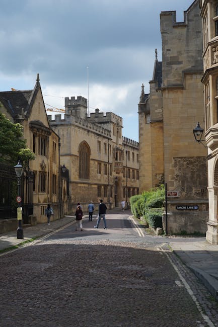 A narrow cobblestone street in a historic city, flanked by beige and light brown stone buildings with Gothic architectural features, including pointed arches and crenellated towers. The street is lined with small black lampposts and has a few pedestrians walking along the sidewalk, some in casual clothing. On the right, a stone building has a small street sign reading 'Magde Lane,' and neatly trimmed green bushes grow beside it. The sky above is partly cloudy with patches of blue visible, casting diffuse natural light on the scene. The setting suggests a European city with a mix of old and well-preserved architecture, typical of an area where private or independent waste disposal services, like those offered by Waste Disposal Merton, might operate for on-site clearance or rubbish removal in historic urban environments.