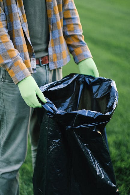A person wearing a green long-sleeve shirt, a checked yellow and blue jacket, beige trousers, and light green gloves is holding open a large black plastic rubbish bag outdoors on a grassy area. The bag is partially filled and appears to be used for waste collection or disposal. The background shows a blurred expanse of green grass, suggesting a park or garden setting. The scene indicates an activity related to waste handling or rubbish removal, which aligns with the services offered by Waste Disposal Merton, emphasizing the importance of proper waste collection through private or independent means rather than municipal services.