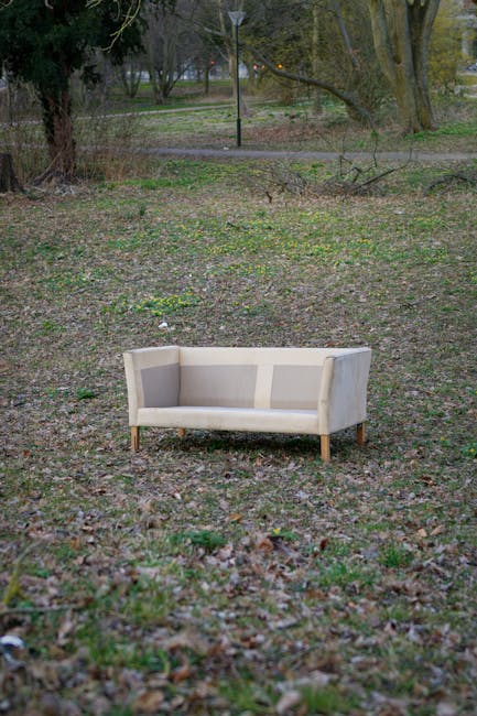 A beige upholstered sofa with wooden legs positioned on a patch of bare ground and sparse grass in an outdoor park area. The sofa appears to be in good condition, with a smooth fabric surface and simple, clean lines. It is centrally located in the image, with trees and a pathway visible in the background. The surrounding environment features natural elements such as fallen leaves, small plants, and a few patches of greenery. In the distance, there is a lamp post and a few more trees, indicating a park setting. The scene is evenly lit, suggesting daytime with natural lighting. The presence of the sofa outdoors implies that it has been placed here for collection or disposal, consistent with services provided by Waste Disposal Merton for private waste handling or alternative rubbish removal. The setting reflects a typical outdoor area where unwanted furniture items might be temporarily placed before collection or disposal, emphasizing the importance of professional rubbish removal services in managing waste collected from residential or communal outdoor spaces.