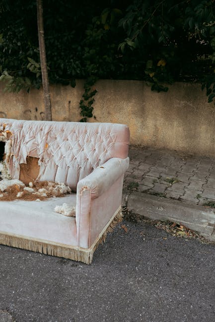 A beige upholstered sofa with wooden legs positioned on a patch of bare ground and sparse grass in an outdoor park area. The sofa appears to be in good condition, with a smooth fabric surface and simple, clean lines. It is centrally located in the image, with trees and a pathway visible in the background. The surrounding environment features natural elements such as fallen leaves, small plants, and a few patches of greenery. In the distance, there is a lamp post and a few more trees, indicating a park setting. The scene is evenly lit, suggesting daytime with natural lighting. The presence of the sofa outdoors implies that it has been placed here for collection or disposal, consistent with services provided by Waste Disposal Merton for private waste handling or alternative rubbish removal. The setting reflects a typical outdoor area where unwanted furniture items might be temporarily placed before collection or disposal, emphasizing the importance of professional rubbish removal services in managing waste collected from residential or communal outdoor spaces.