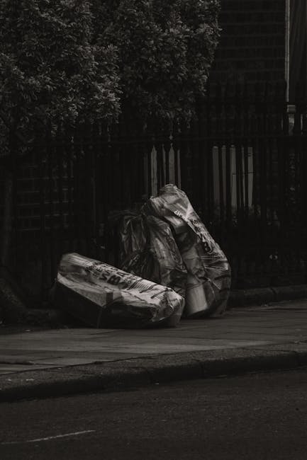 Three large black garbage bags filled with waste material are placed on the pavement next to a dark metal fence, with a background of leafy trees and a brick building. The bags are made of thick plastic, showing some creases and slight reflections from ambient light. They are positioned in a loose cluster, leaning against each other, with one partially resting on the curb and others slightly upright on the sidewalk. The scene appears to be in an urban or suburban street, where private rubbish disposal, such as this, might be managed independently of local council services. The photograph is in black and white, emphasizing the textures of the plastic bags and the surrounding environment. The bags are situated next to the sidewalk, close to a mature hedge on the left and a high brick wall or building on the right, indicating a typical street scene where waste has been collected in preparation for removal by a professional waste disposal service like Waste Disposal Merton.