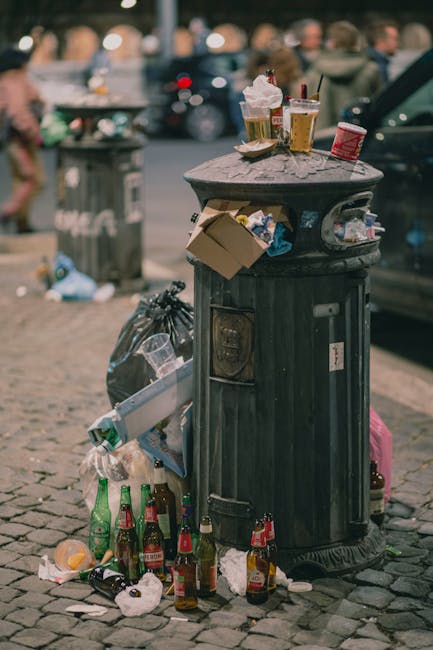 A black, cylindrical public rubbish bin with a domed, textured lid situated on a stone-paved urban street, surrounded by litter including empty bottles, crumpled paper, and discarded food containers at its base. Several paper cups and drink cans are placed on top of the lid, some with used straws and ice residue, indicating recent consumption. A small cardboard box and a plastic bag are draped over the side of the bin, with additional trash strewn on the ground nearby. In the background, blurred street scene elements such as pedestrians, parked cars, and a pedestrian crossing signal are visible, suggesting a busy city environment during daylight hours. Waste Disposal Merton provides services to manage rubbish removal and private waste disposal, ensuring proper handling of such clutter in urban settings, though the scene depicted highlights the importance of timely rubbish collection to maintain cleanliness.
